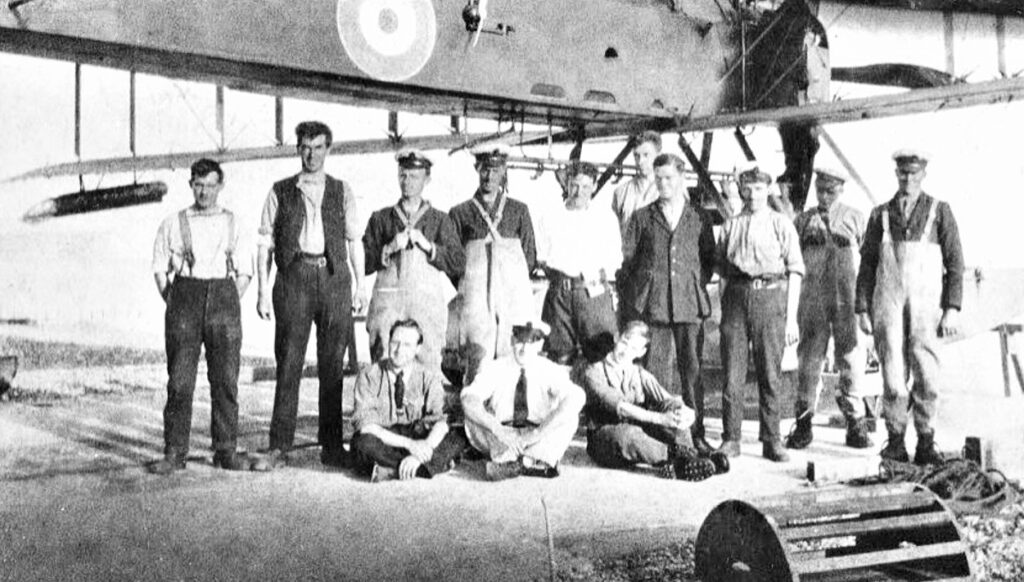 A black 7 white photograph of 13 men, 10 standing and 3 sitting cross-legged under a seaplane.